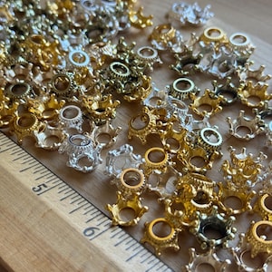 May include: An assortment of miniature crown-shaped metal findings in gold, silver, and bronze hues. The findings are scattered on a wooden surface next to a ruler, highlighting their detailed design and diverse finishes.