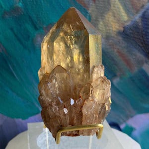Puede incluir: Un grupo de cristales de cuarzo de color marrón dorado. El cristal principal es un prisma alto de seis lados con una parte superior puntiaguda. Cristales más pequeños rodean la base. El cuarzo está montado sobre un soporte de metal dorado, con un fondo abstracto colorido.