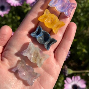 Puede incluir: Cinco tallas de cristal en forma de caramelo de colores en una mano. Los cristales son en tonos morado, amarillo, azul y transparente. El fondo incluye hierba verde y flores moradas.