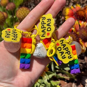 May include: A collection of keychains featuring colorful rainbow-colored building blocks and yellow charms. Each charm has the words "Happy Day" printed on it. The keychains are held in a hand against a blurred background of flowers.