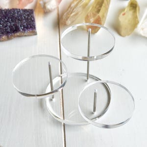 Puede incluir: Un expositor de acrílico de tres niveles con plataformas circulares transparentes y varillas de metal plateado. El expositor está diseñado para sostener objetos pequeños, como cristales o joyas.