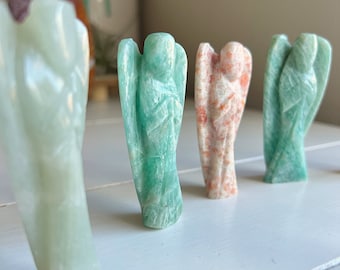 May include: Five carved angel figurines made of green and orange stone. The figurines are arranged in a row on a white surface.