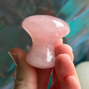 Puede incluir: Una herramienta de masaje facial en forma de hongo de cuarzo rosa. La herramienta se sostiene en una mano, mostrando su superficie lisa y pulida. El fondo es una mezcla borrosa de verde azulado y azul.