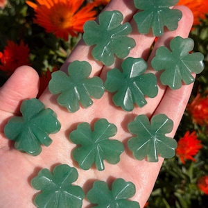 Könnte beinhalten: Eine Sammlung grüner Aventurin-Kleeblatt-Anhänger. Jeder Anhänger ist in Form eines vierblättrigen Kleeblatts mit detaillierten Adern geschnitzt. Die Anhänger werden in einer Hand gehalten, mit orangefarbenen Blumen im Hintergrund.