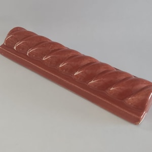 May include: A terracotta-colored ceramic tile with a rope-like design. The tile is slightly curved and has a glossy finish.