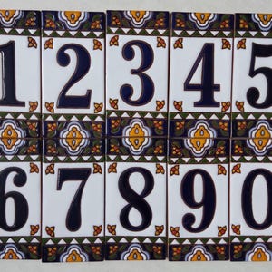 May include: A set of ten decorative ceramic tiles, each displaying a different number from 0 to 9. The numbers are in a bold, dark blue font against a white background. Each tile features a decorative border with floral and geometric patterns in blue, yellow, and green.