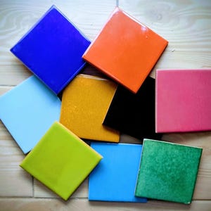 May include: A collection of square ceramic tiles in various colors, including blue, orange, pink, green, and yellow. The tiles are arranged on a light wood surface, showcasing their glossy finish and vibrant hues. Each tile appears to be approximately 4 inches by 4 inches.