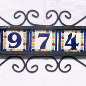 May include: Decorative address plaque featuring the numbers 69742. The numbers are in a bold blue font on a white background with floral accents. The plaque is framed by a black wrought iron design.