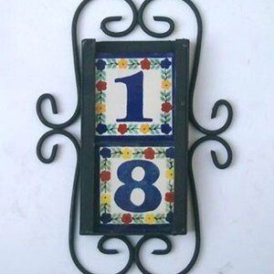 May include: A black metal frame with scrolled details holds two ceramic tiles with the numbers 1 and 8. The tiles are decorated with a floral pattern in shades of blue, yellow, red, and green.