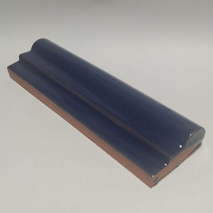 May include: A navy blue ceramic tile with a glossy finish. The tile has a rounded top edge and a rectangular base with a reddish-brown color. The tile is designed for architectural or decorative use.