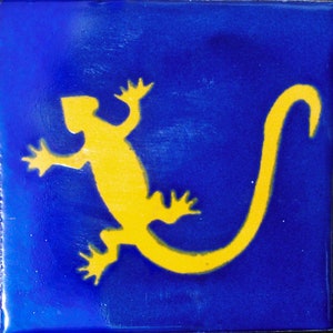 May include: Square blue tile with a yellow lizard design. The lizard is stylized with a long tail and outstretched limbs. The tile has a glossy finish and a smooth surface.