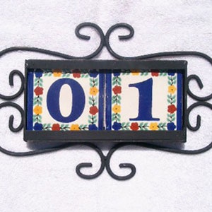May include: Decorative house number sign featuring the numbers "0" and "1" on ceramic tiles. The tiles are framed in a black metal holder with decorative scrollwork. The tiles have a white background with blue numbers and floral accents.