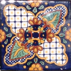 May include: A square ceramic tile with a blue background and a white, yellow, and red floral pattern. The tile has a geometric design with a central motif of a stylized flower.
