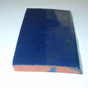 May include: A rectangular ceramic tile featuring a deep, glossy navy blue glazed finish on the top surface. The side profile reveals a natural, unglazed terracotta-colored clay base with a slightly curved edge along one of the long sides.