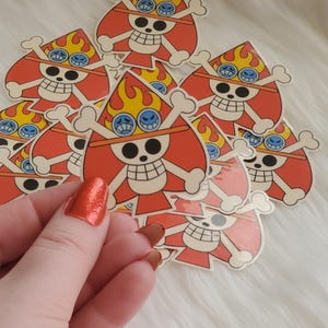 Könnte beinhalten: Eine Sammlung roter, orangefarbener und gelber Aufkleber mit einem Totenkopf- und gekreuzten Knochen-Design mit Flammen und einer Brille. Die Aufkleber werden in einer Hand vor einem weißen, flauschigen Hintergrund gehalten.