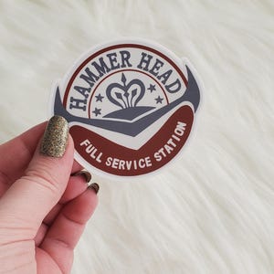 以下が含まれることがあります： 赤とグレーのデザインの白いステッカー。ステッカーには「Hammer Head Full Service Station」とスタイリッシュな王冠と星が書かれています。