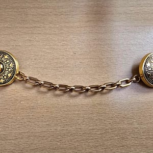 May include: A gold-toned metal cardigan clip featuring two circular fasteners connected by a short chain. Each fastener has a raised, intricate, dark-accented floral pattern surrounding a smooth, polished, convex center dome.