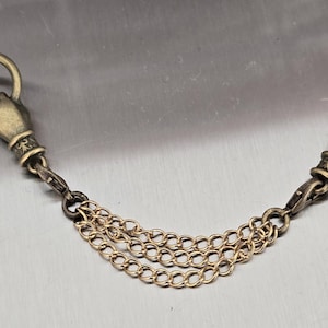 May include: An antique gold-coloured chain featuring two hand-shaped charms, each holding a ring. The chain comprises three parallel strands. The hand charms are detailed with intricate designs. Likely a decorative accessory.