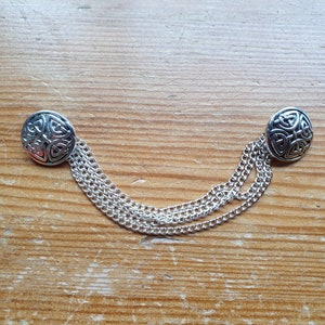May include: A silver chain with two round, silver Celtic knot buttons. The chain is made of multiple strands and the buttons are connected to the chain.