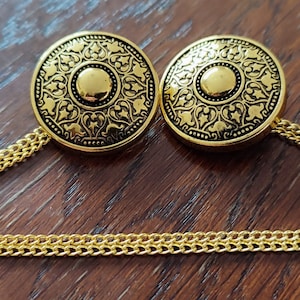 May include: A pair of gold-tone metal collar stays with a decorative chain connecting them. The stays feature a circular design with intricate floral patterns.