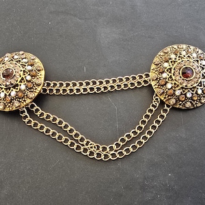 May include: A vintage gold-toned brooch with two circular, ornate medallions connected by double chains. Each medallion features a central dark red stone surrounded by smaller white, brown, and clear crystals. The brooch is a decorative accessory.
