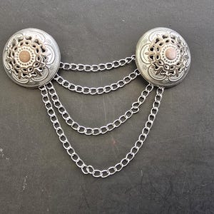 May include: Silver-coloured metal brooch featuring two circular decorative elements linked by three chains. Each circular element has an intricate floral design with a central, light brown accent. The brooch is set against a dark background.