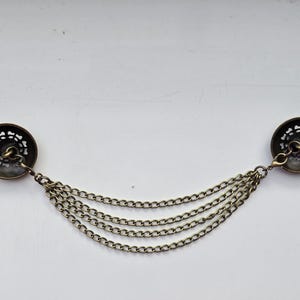May include: Antique brass-toned collar clip with two round, decorative button-like clasps connected by four strands of delicate chain. The clasps have a floral-like cutout design.