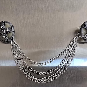 May include: Silver-toned brooch with two round, decorative ends connected by multiple strands of chain. Each end features a cross design with small, clear embellishments. The brooch has an antique or vintage aesthetic.