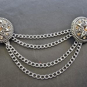 May include: Silver-toned brooch with two round, ornate medallions connected by three strands of chain. Each medallion features a floral design with amber-colored accents, set against a textured background. A decorative accessory.