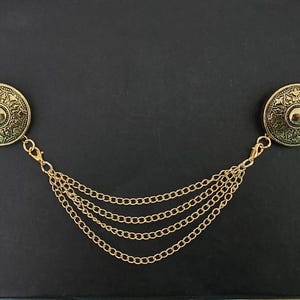Può includere: Una clip per cardigan in metallo dorato con due bottoni circolari decorati con intricati motivi in rilievo. I bottoni sono collegati da quattro delicate catenelle dorate drappeggiate che pendono in una formazione curva e stratificata.