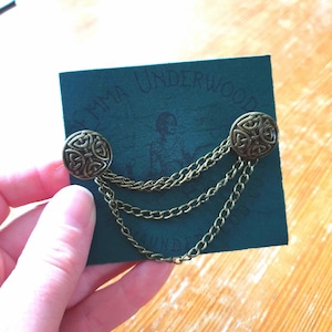 Pode incluir: Um par de broches de nó celta de metal dourado conectadas por uma corrente. As broches estão em um cartão verde com o texto "Emma Underwood" impresso nele.