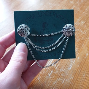 Könnte beinhalten: Eine silberne Kette mit zwei Knöpfen im keltischen Knotendesign auf einer grünen Karte mit dem Text "Emma Underwood".