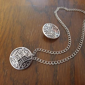 May include: A silver chain necklace with a silver pendant featuring a circular design with intricate details. The pendant is attached to the chain with a silver clasp.