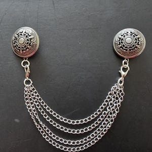 Könnte beinhalten: Silberfarbene Brosche mit zwei kreisförmigen Dekorationselementen, die durch mehrere Ketten verbunden sind. Jedes kreisförmige Element weist ein aufwendiges Blumendesign auf. Die Brosche ist zum Anbringen an Kleidung konzipiert.
