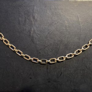 May include: A gold-toned chain with two circular, brass-colored clasps. The clasps are connected by a chain of small, rectangular links. The background is a dark, textured surface.