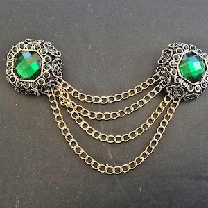 May include: A decorative brooch with two square-shaped elements, each set with a large, faceted emerald-green gemstone. Multiple strands of gold-coloured chain connect the elements. The brooch has an antique, vintage design, suitable for formal wear or adding flair.