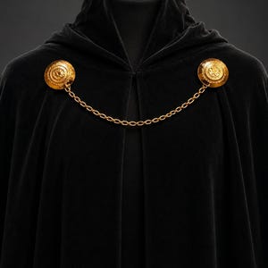 May include: A black velvet cloak with a hood, fastened by two gold-colored circular clasps connected by a chain. The clasps have an ornate design. The cloak is displayed on a mannequin.