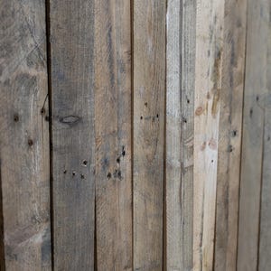 May include: Close-up of a wooden wall made of vertical planks. The wood is a mix of light and dark brown colours, with some knots and imperfections. The planks are arranged in a tight pattern, creating a rustic and natural look.