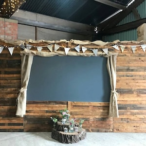 May include: A rustic wooden backdrop with a hessian curtain on each side. A large chalkboard is centred on the wall. A string of lights and a banner of white flags hang above the chalkboard. A small arrangement of flowers and candles sits in front of the backdrop.