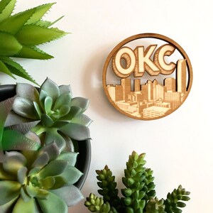 May include: A wooden round ornament with a cutout of the Oklahoma City skyline and the text "OKC" in a retro font.