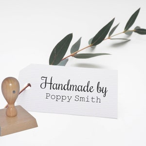 May include: A wooden stamp with a white label that reads "Handmade by Poppy Smith". The stamp is on a white surface with a sprig of green eucalyptus leaves.
