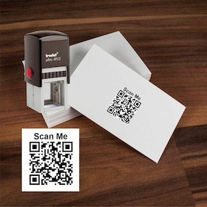 May include: A black and white QR code with the text "Scan Me" printed on a white business card. A black self-inking stamp with the text "trodat printy 4923" is in the background.