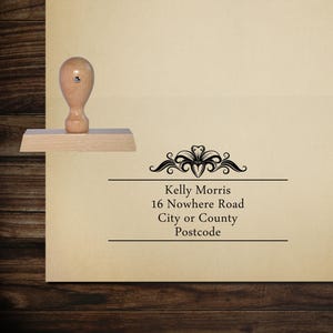 May include: Wooden self-inking address stamp with a decorative design. The stamp reads "Kelly Morris, 16 Nowhere Road, City or County, Postcode".