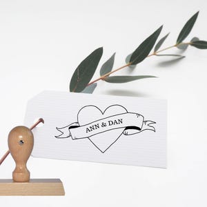 May include: A white card with a black heart and banner that reads "ANN & DAN". The card is sitting on a wooden base with a wooden stamp handle.