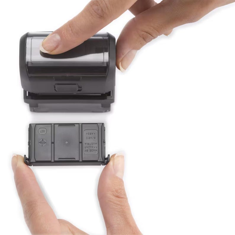 Op de afbeelding: Een zwarte zelf-inktende stempel met een transparant venster en een vervangbare inktcartridge. De cartridge is ge&euml;tiketteerd met "MADE BY TRODAT AUSTRIA 12/17-2  4483".
