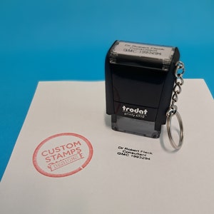May include: A black self-inking stamp with a white label that reads "Dr Robert Fleck Consultant GMC 1993294". The stamp is attached to a silver keychain. A red circle stamp with the text "CUSTOM STAMPS DESIGNS" is visible on a white surface.