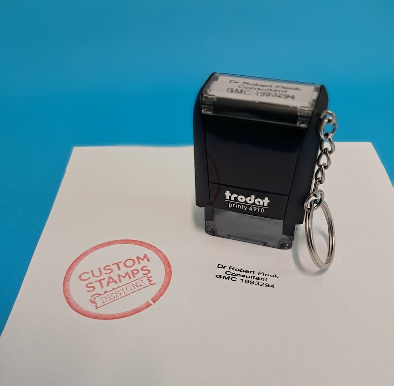 May include: A black self-inking stamp with a white label that reads "Dr Robert Fleck Consultant GMC 1993294". The stamp is attached to a silver keychain. A red circle stamp with the text "CUSTOM STAMPS DESIGNS" is visible on a white surface.