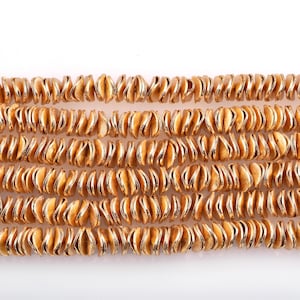 Peut inclure: Un brin de perles en métal doré, chaque perle est en forme de petite feuille enroulée. Les perles sont disposées en rangée, créant un effet chatoyant et métallique.