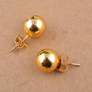 Könnte beinhalten: Ein Paar goldfarbene Kugel-Ohrstecker. Die Ohrringe haben ein schlichtes Design und sind aus glänzendem Metall gefertigt.