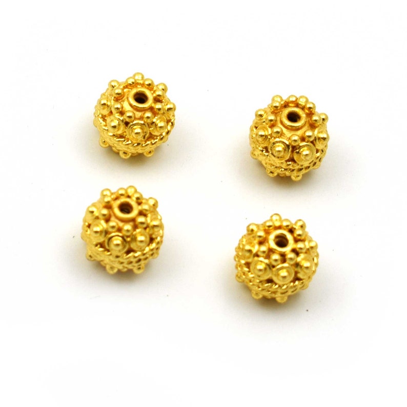 May include: Four gold-colored, spherical beads with a textured surface. Each bead has a central hole and is covered in small, raised circular details. The beads are evenly spaced against a white background, showcasing their intricate design.